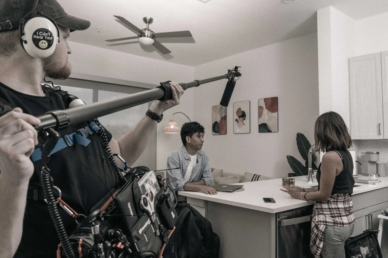 A sound recordist holding a boom microphone above two performers during a kitchen scene in Recovery.